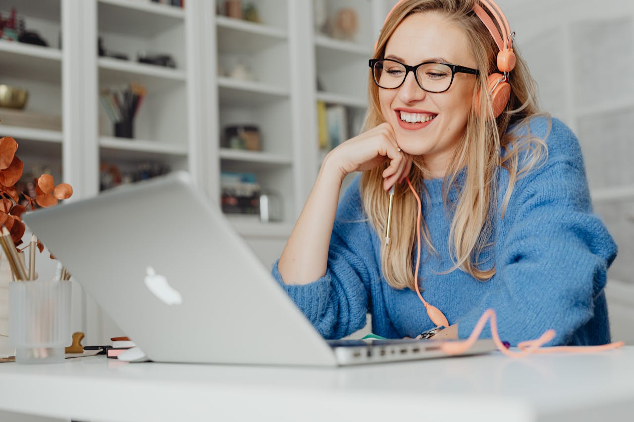Smiling woman wearing headphones and glasses, working on a laptop in a bright indoor setting.