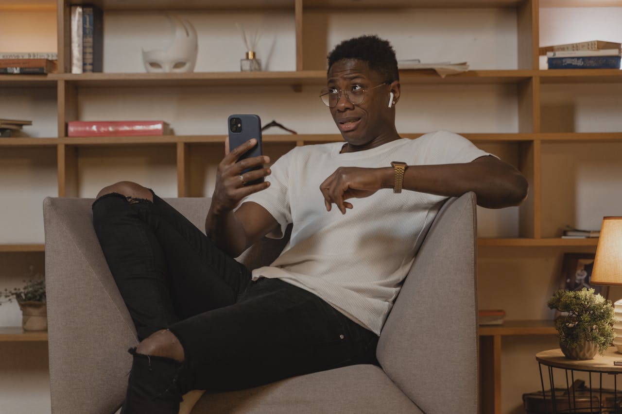 Young man in casual attire chatting on smartphone while lounging on a chair indoors.