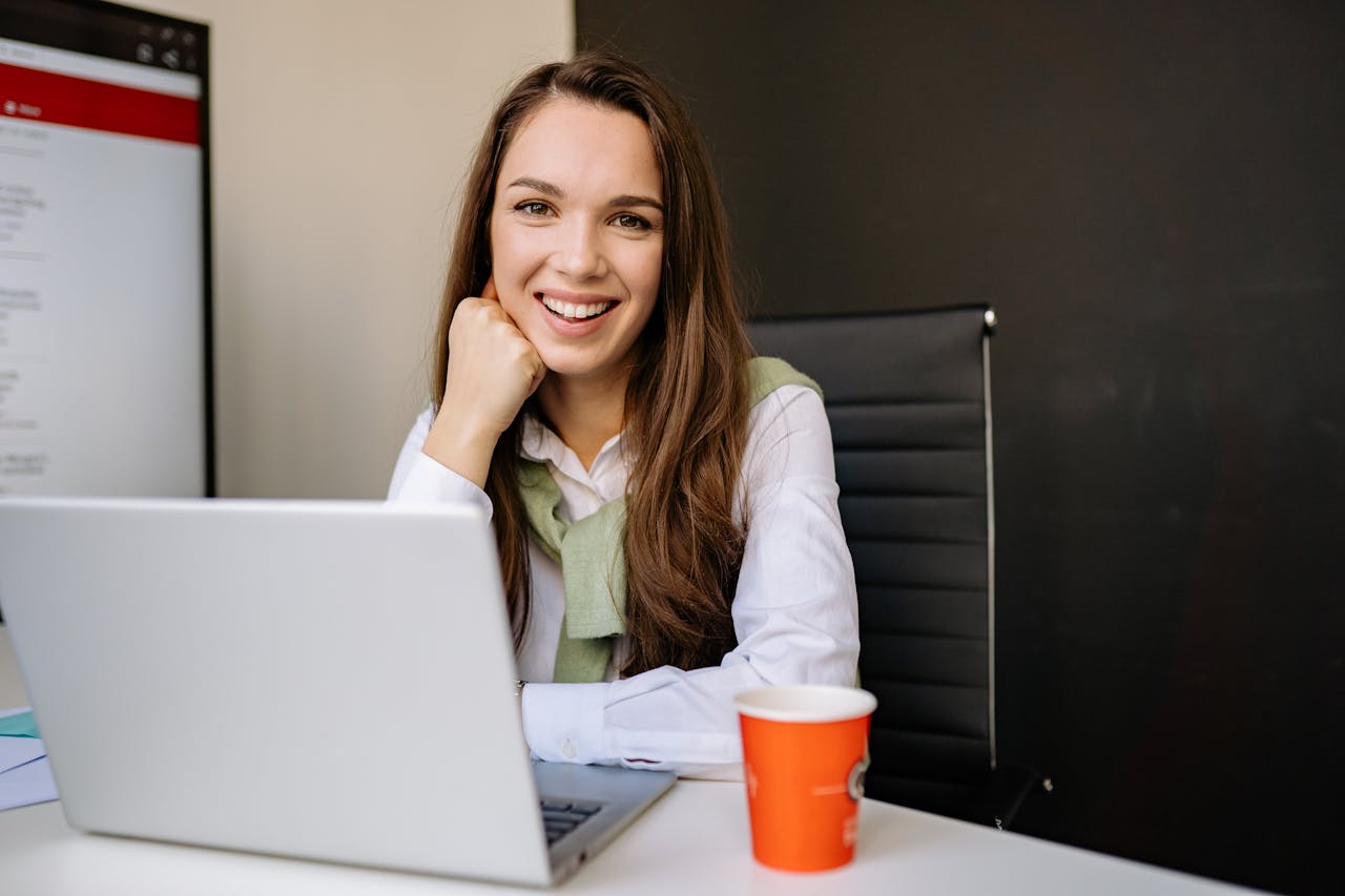 Businesswoman sitting at desk in office, smiling at camera with a laptop and coffee cup.